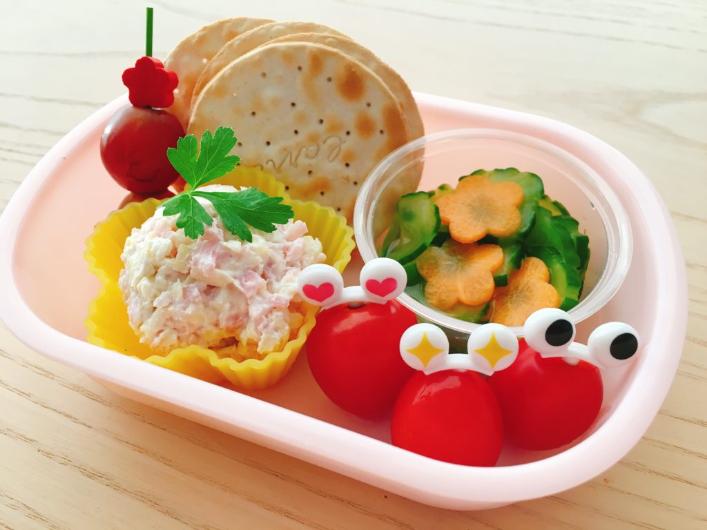Ham spread, crackers, carrot and cucumber salad, and cherry tomatoes are dressed up for this young child's bento.