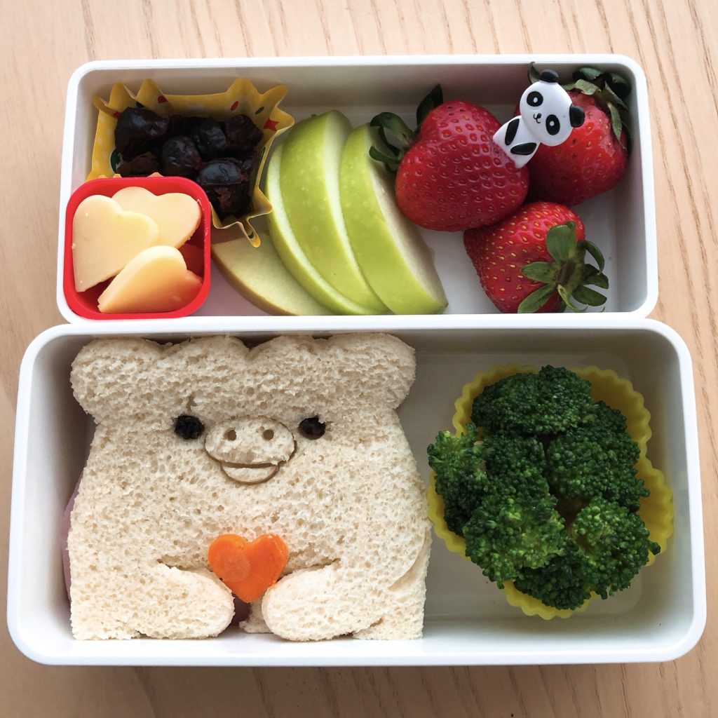 A sandwich cutter is used for a ham and cheese bear sandwich. Cheese hearts , strawberries, apples, broccoli and raisins complete the bento box.