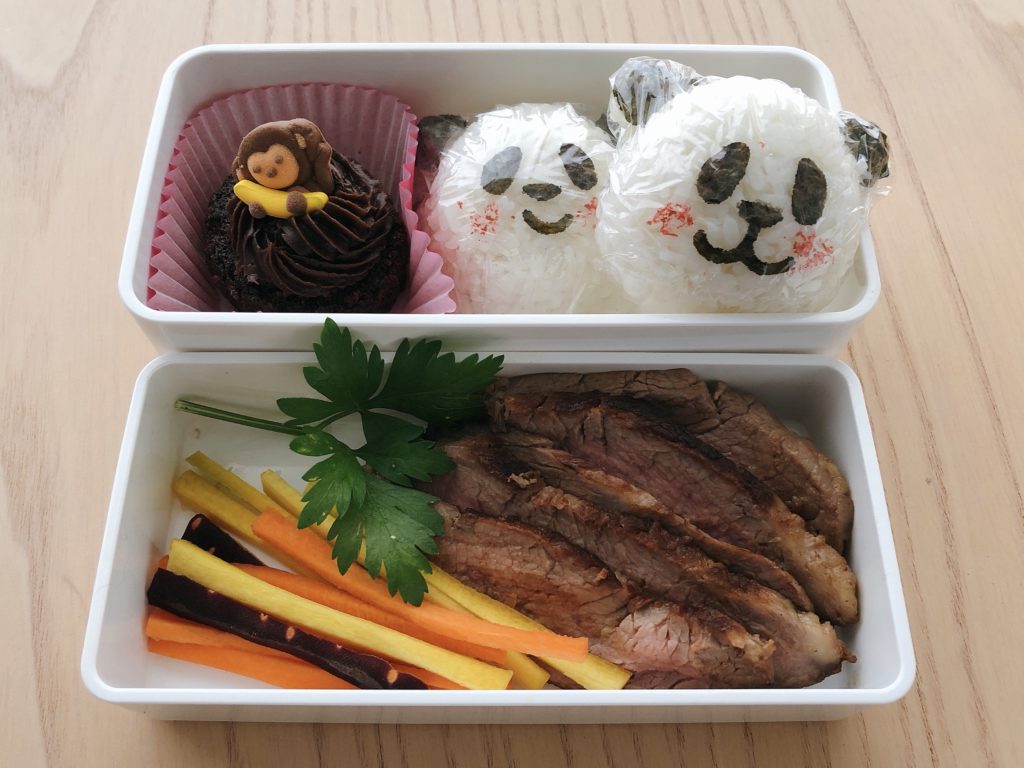 Steak slices, carrot sticks, rice balls shaped like pandas and a mini chocolate cupcake make a birthday bento lunch.