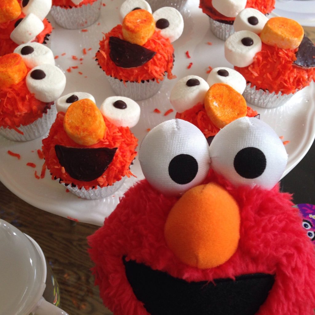 Elmo stuffed toy is surrounded by Elmo cupcakes. These cupcakes are made with marshmallows, coconut, and marzipan-covered chocolate.
