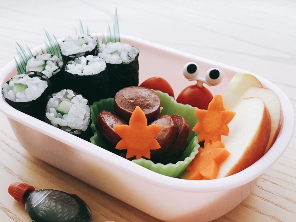Sushi you make at home can be turned into a bento lunch to take to school or work. This child's lunch includes teriyaki hot dogs, cherry tomatoes, and apple slices.