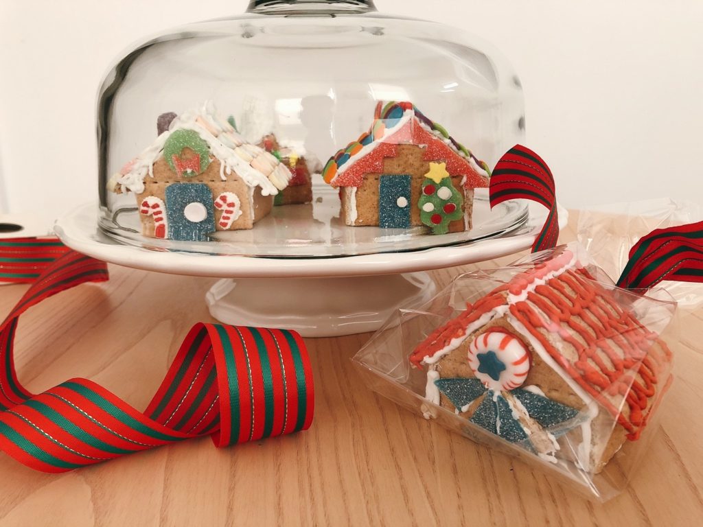 Simple gingerbread houses are stored under a cake dome. Another house is packaged in a cellophane bag for gift-giving.