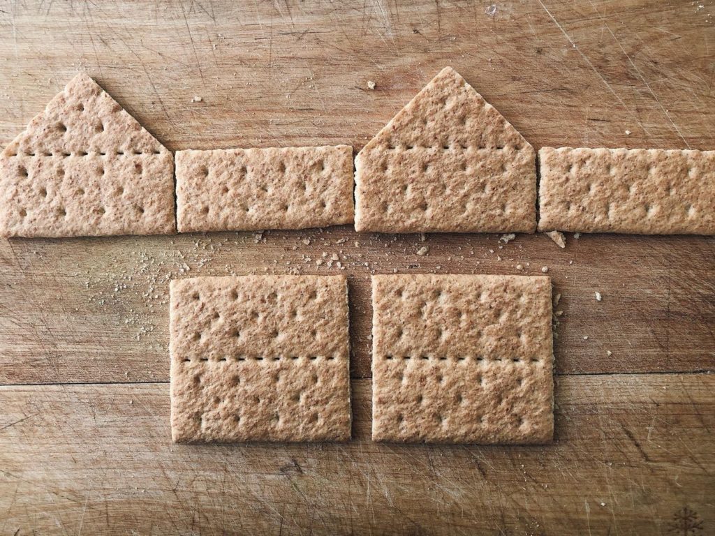 How five graham crackers make a house: two whole crackers are trimmed to peaks to form the front and back walls of the house, one cracker is cut in half for the two side walls, and two crackers are left whole for the roof.