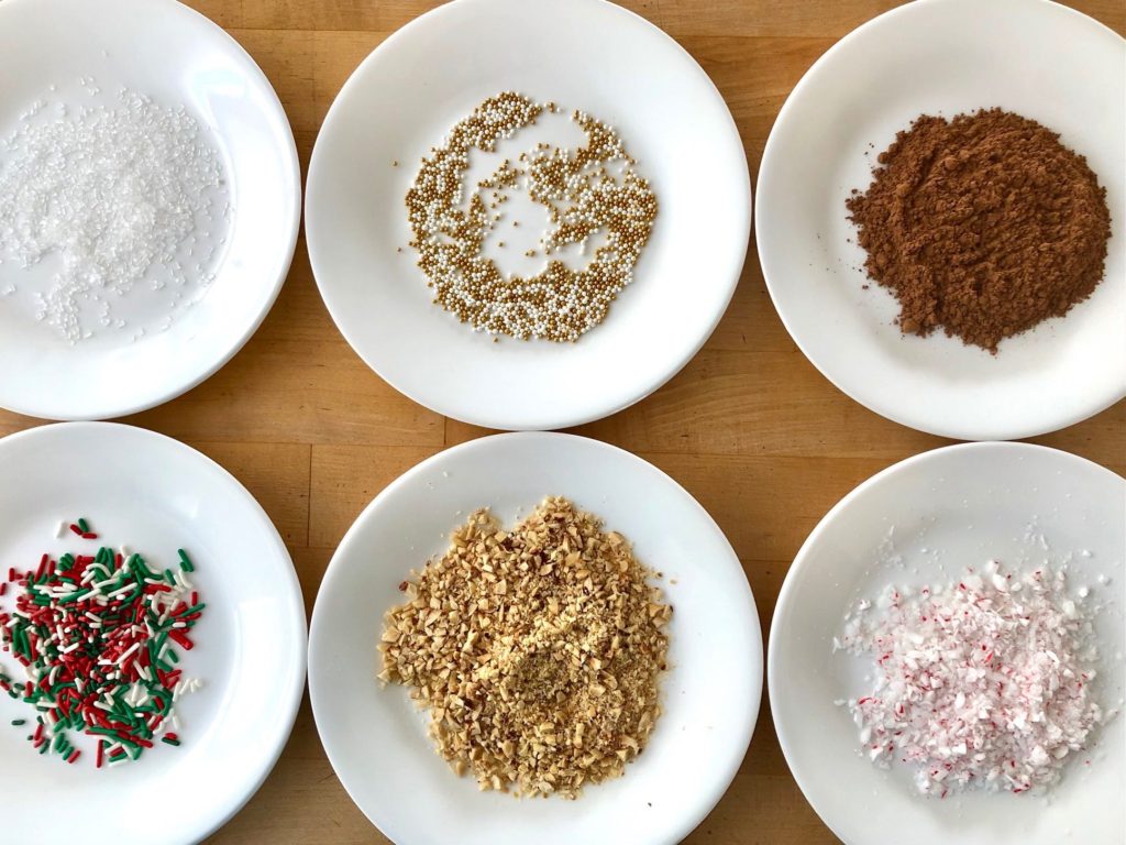 A variety of coatings--from hazelnuts, to cocoa powder, to candy cane and sprinkles--are laid out on plates.