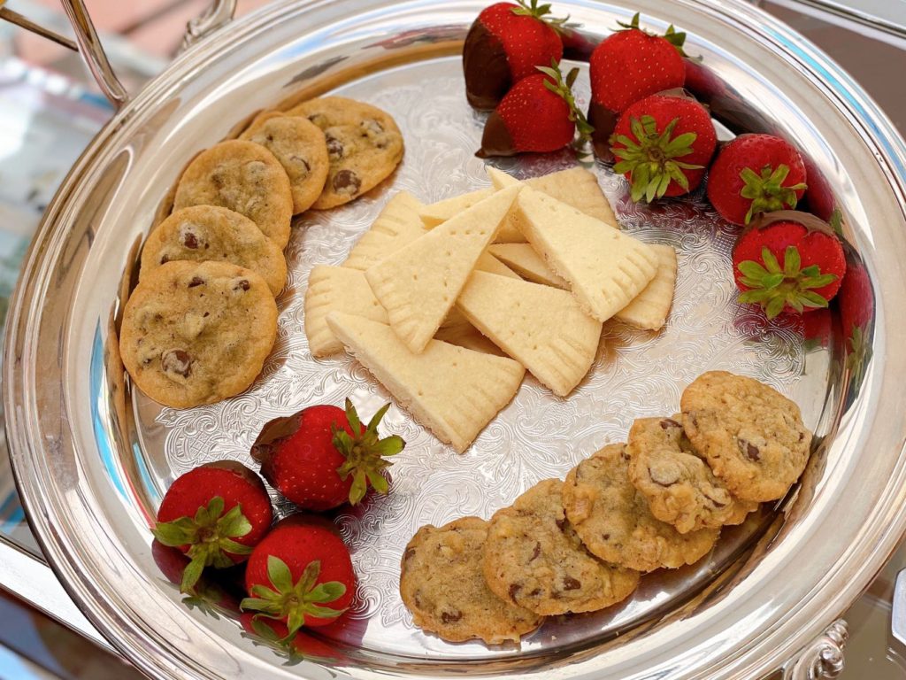 A display of sweets on a silver tray: mini chocolate chip cookies, chocolate-dipped strawberries, and shortbread wedges.
