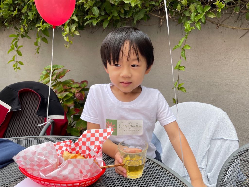 Child sits at a table having a snack at the Camp Grandma summer party.
