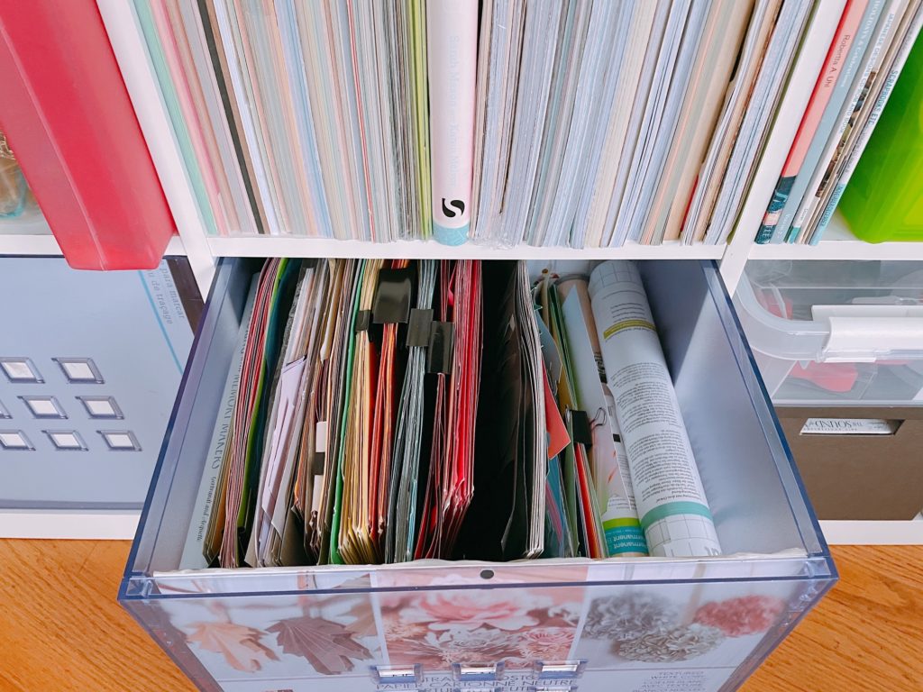 Cardstock is shelved like books; cardstock scraps are grouped by color and pinned together with binder clips, then stored in a plastic bin.