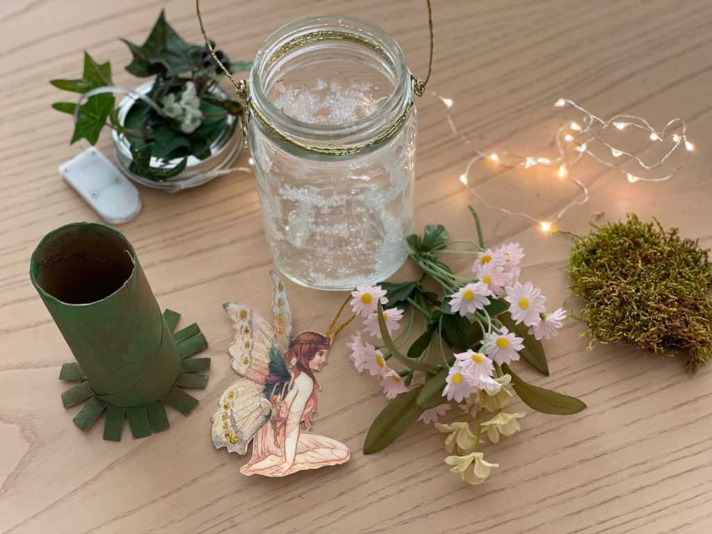 Some of the materials for the fairy lantern project: mason jar with glitter, toilet paper roll, fairy image, artificial flowers and leaves, LED fairy lights, and dried moss.