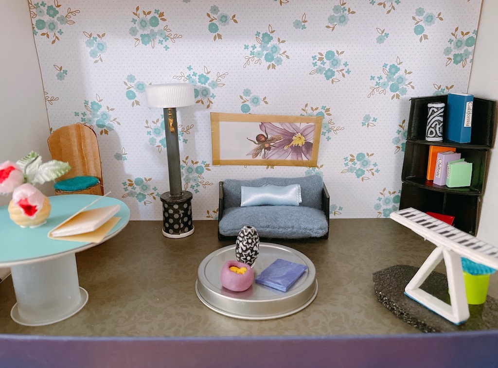 The living room features shelves made from pizza box corners, a table made from a ribbon spool, and a lamp made from a crayon and bottle cap.