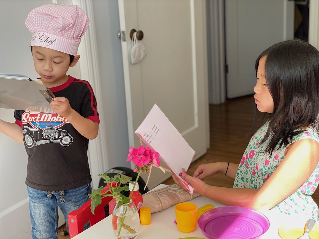 Child ordering food, second chiding checking off the order. They're playing the restaurant game, best game for learning and having fun.
