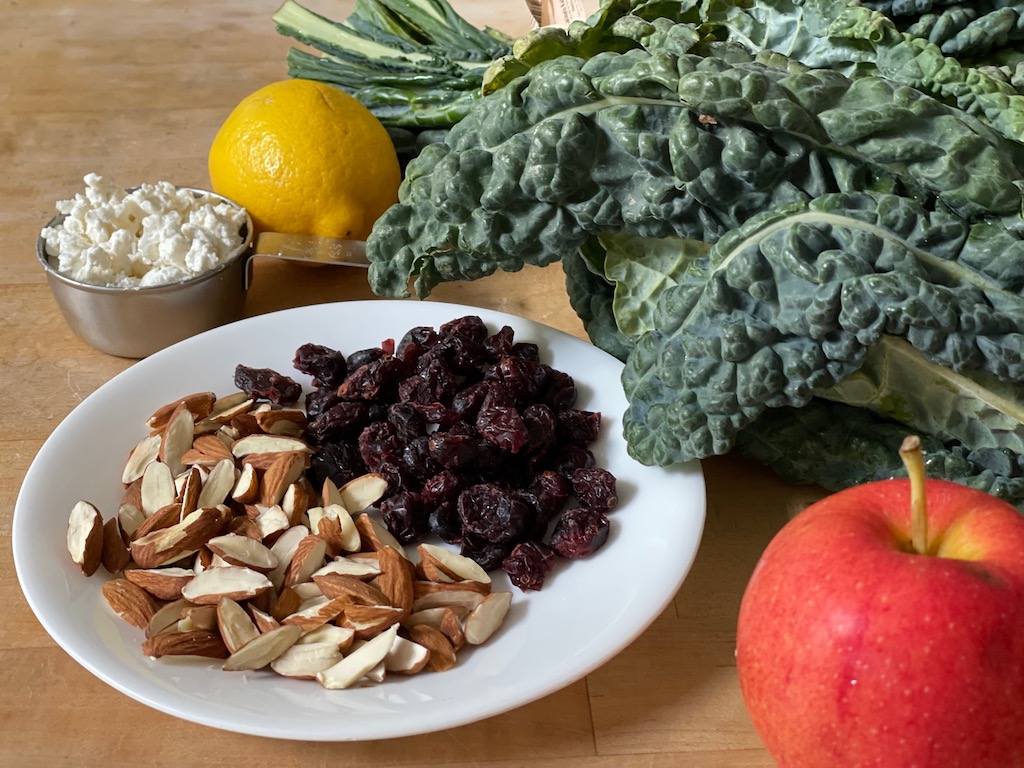 Ingredients for a healthy salad: almonds, dried cranberries, feta cheese, apple, lemon juice, and kale.