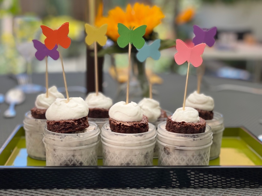 Mini mason jars have a layer of brownie, ice cream, and another brownie, with a topping of whipped cream. The dessert is decorated with paper punch butterflies on skewers.