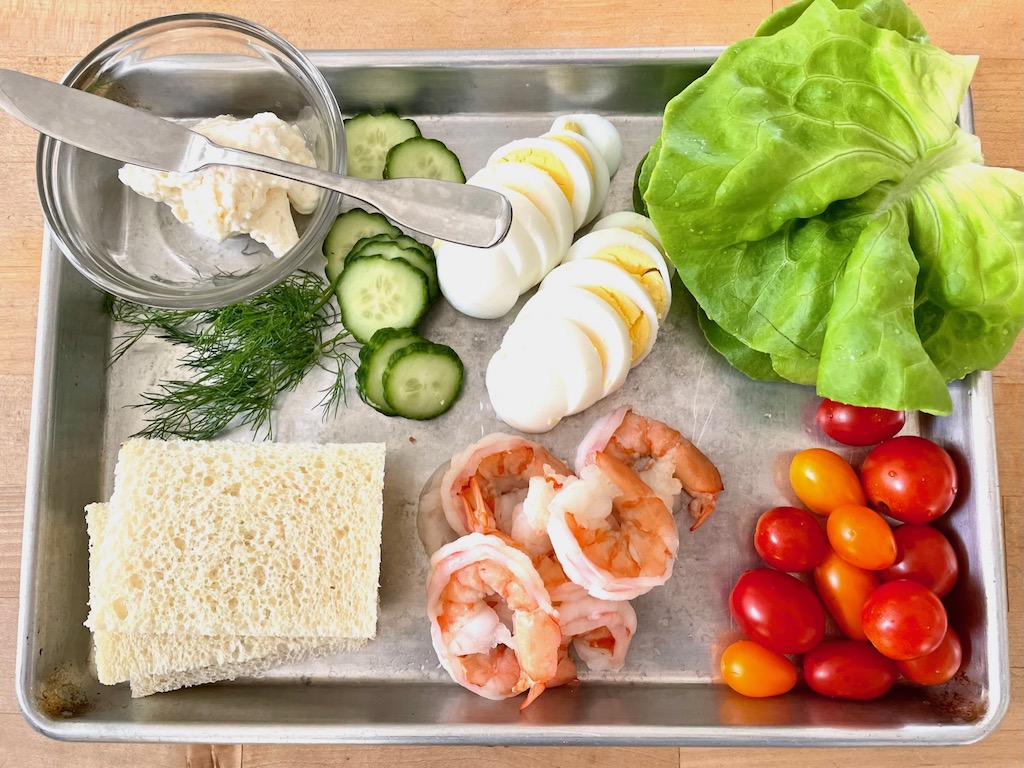 Mayonnaise, dill, cucumber, eggs, lettuce, prawns, and cherry tomatoes are prepped to make into open-faced sandwiches.