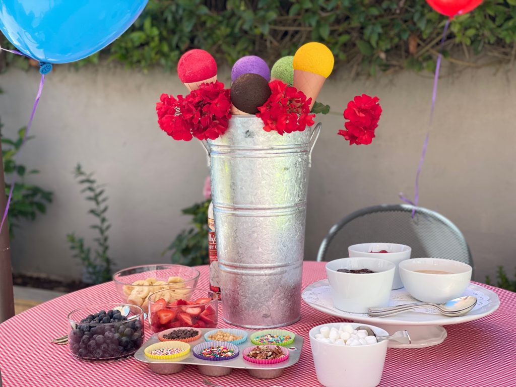 Set up for an ice cream party: fruits in bowls, sprinkles in muffin tins, and sauces on a pedestal cake plate.