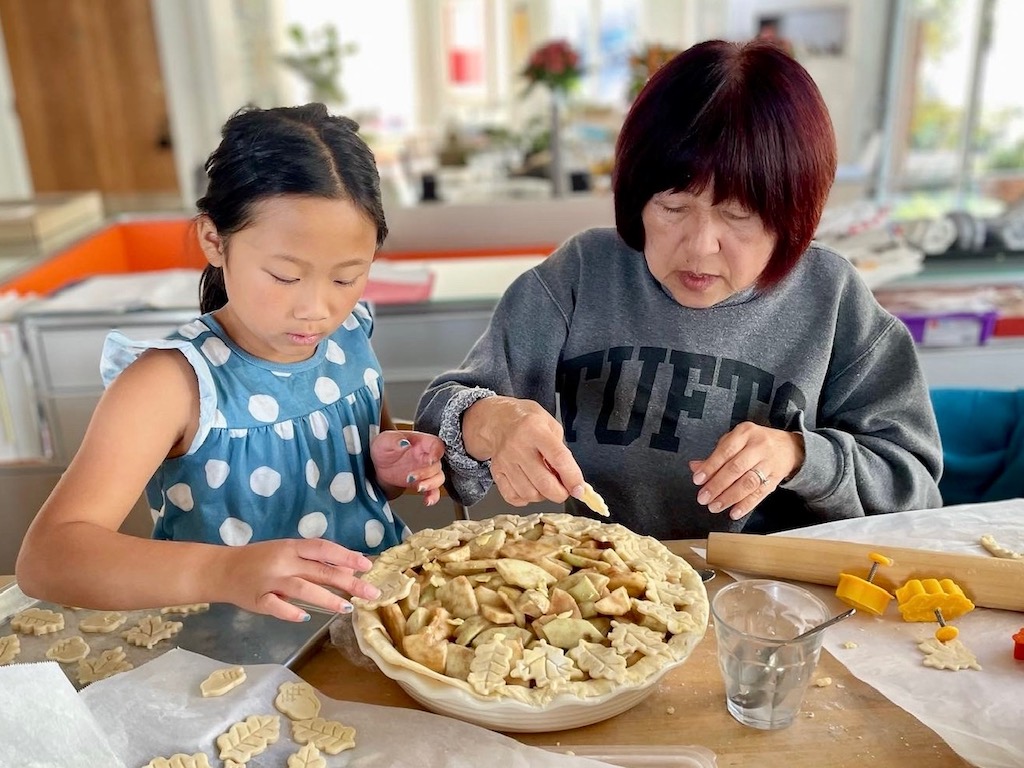 Child and grandma arranging pie crust leaves on an apple pie.