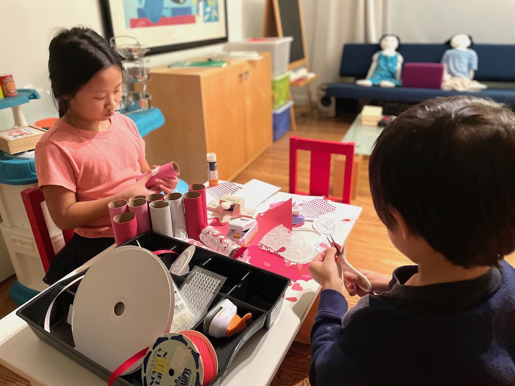 Two children cutting and pasting decorative elements on painted toilet paper tubes.