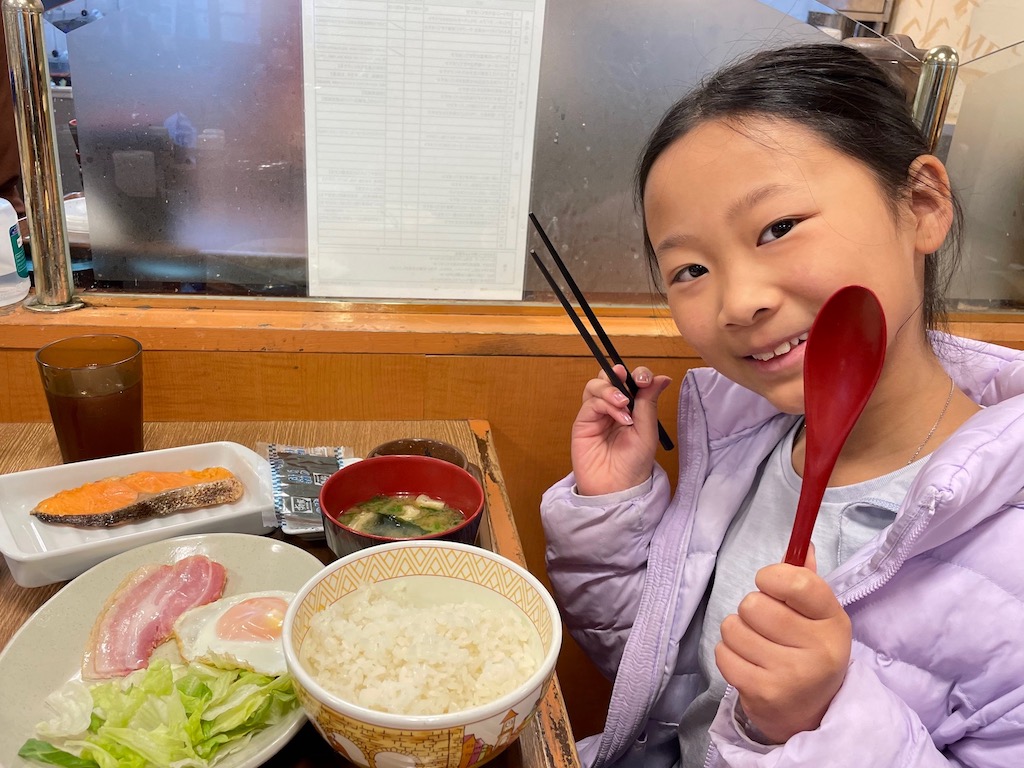 When traveling with kids, be flexible about meals. A child enjoys a Japanese breakfast with bacon and eggs, grilled salmon, miso soup, and rice.