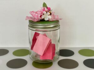 Jar decorated with artificial flowers, holds children's sentiments about mom and promises for services, written on strips of paper and tucked inside.