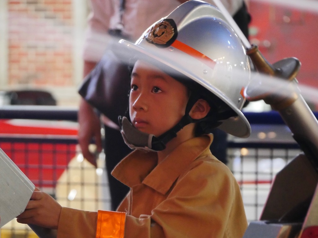 A child if full firefighter's uniform helps to extinguish the "fire" with a hose.