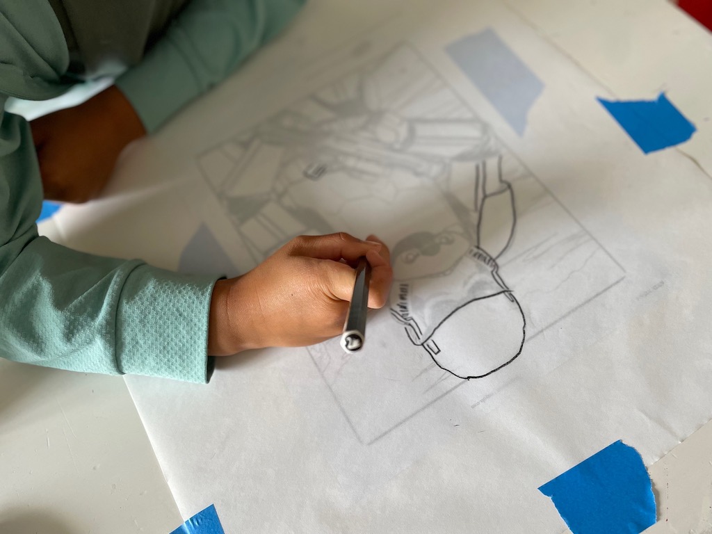 First tracing: child traces the artwork with a soft graphite pencil. The artwork and tracing paper are held in place with painter's tape to prevent slipping.
