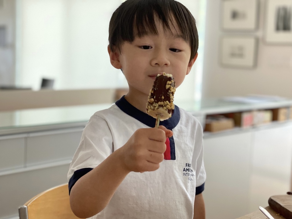 Cool off with a healthy frozen treat. Child eats a banana pop, frozen banana on a stick, covered in chocolate.