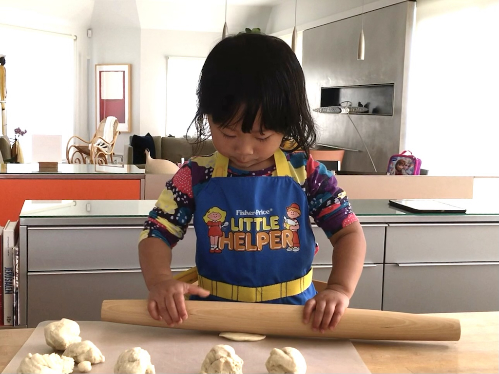 Three-year-old child rolls out pizza dough for mini pizzas.