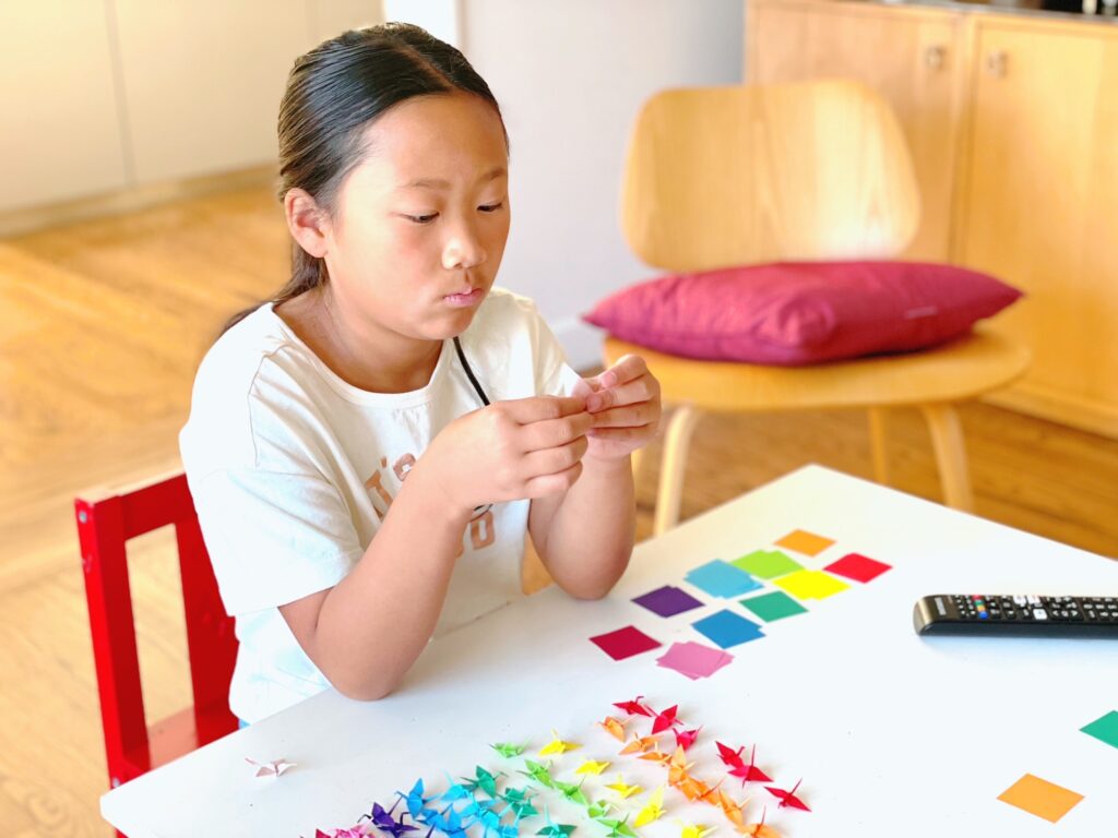 Child folding mini origami cranes, with rows of finished cranes arranged by color.