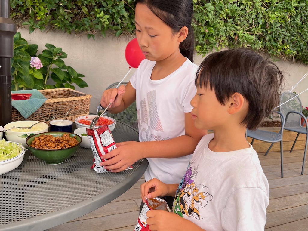 Kids fill their chips bag with taco fixings to eat from the bag.