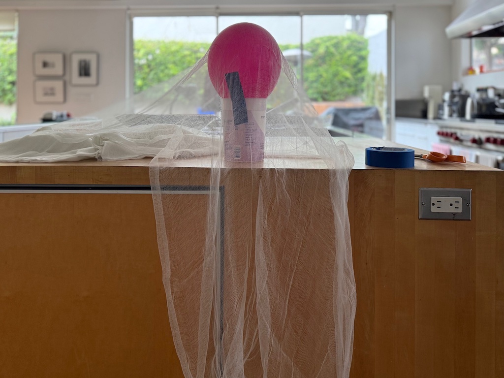 Balloon is taped to a large yogurt container. The cheesecloth is draped over the balloon to determine how long to cut the cheesecloth.