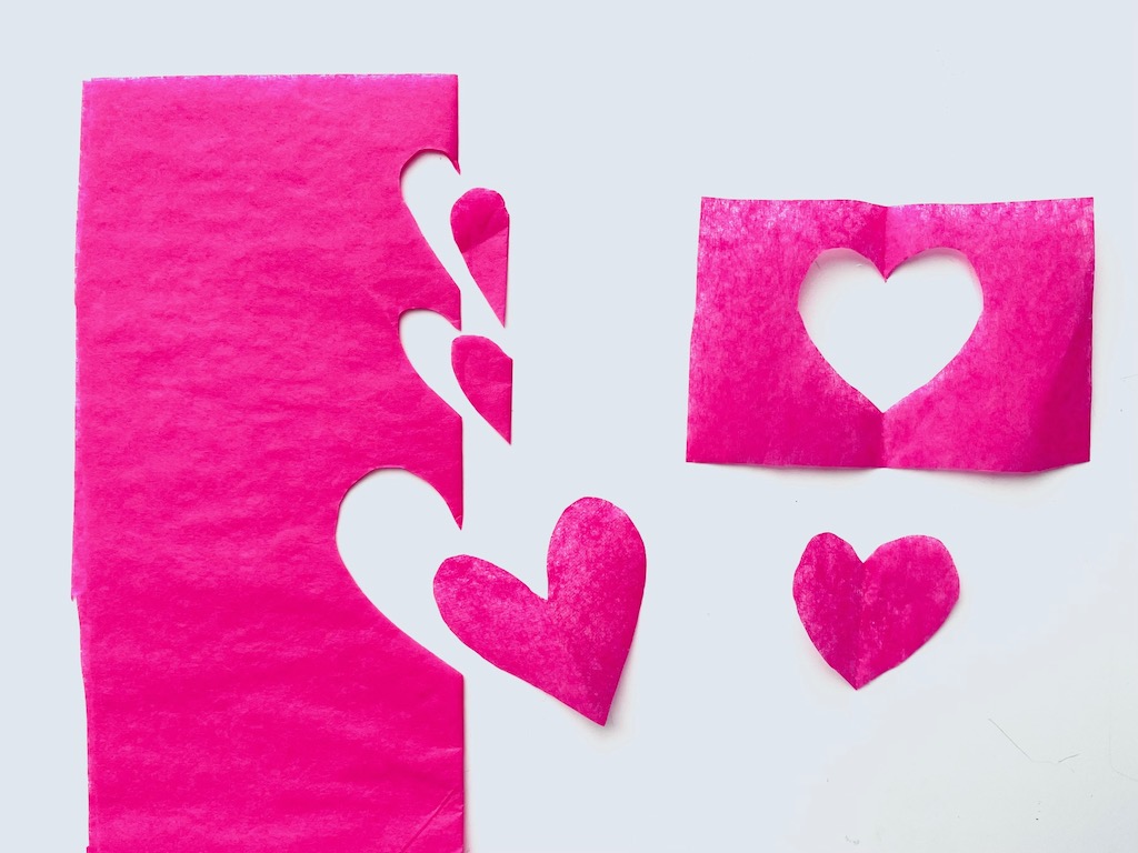 To make tissue paper hearts, fold tissue paper in half and cut out half heart shapes along the fold, freehand. Open the fold for the complete heart.