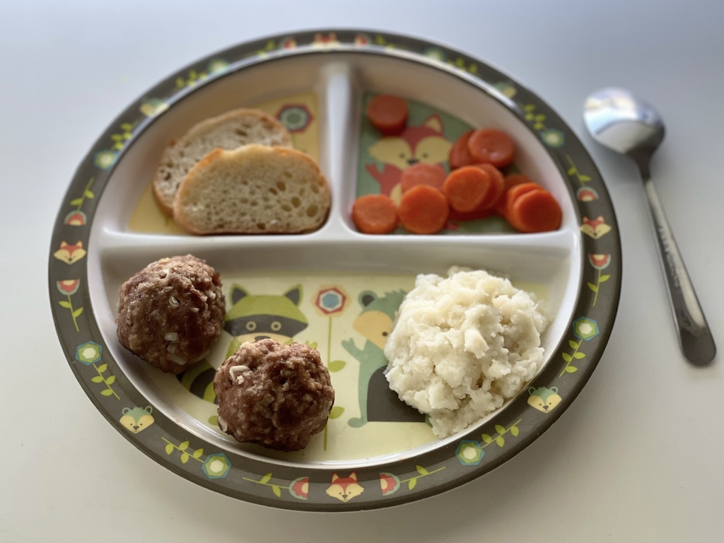 A child's meatball dinner: two meatballs, mashed potatoes, buttered carrots, and baguette slices. 