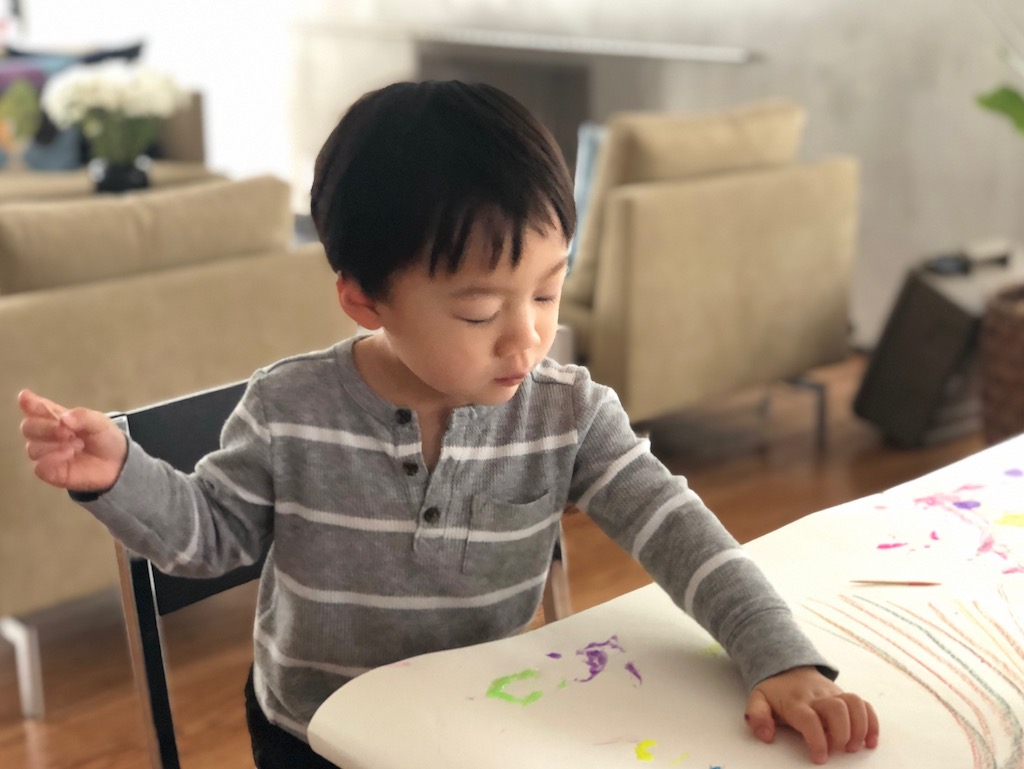 Child sits at a paper-covered table and is given crayons and art tools to draw with.