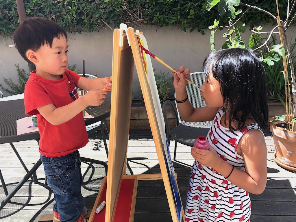 Two children paint at an easel outdoors.