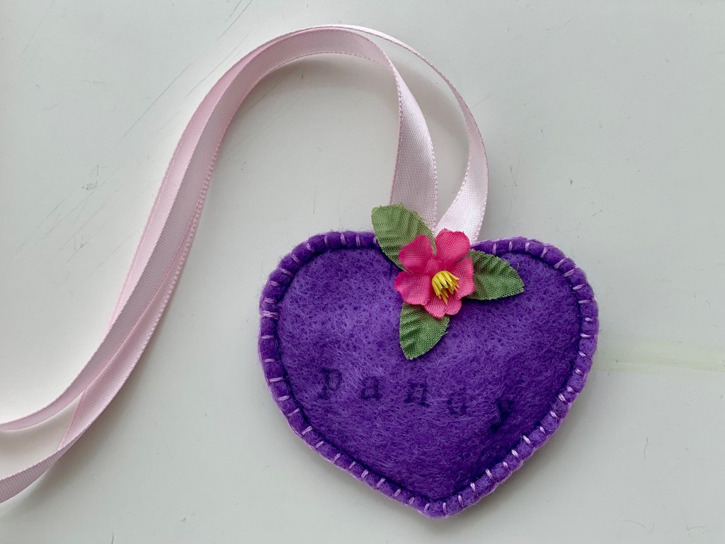 The finished air tag holder is made with felt, stamped with the stuffie's name, and decorated with an artificial flower. Satin ribbons are used to tie the air tag holder around the neck of the stuffie.