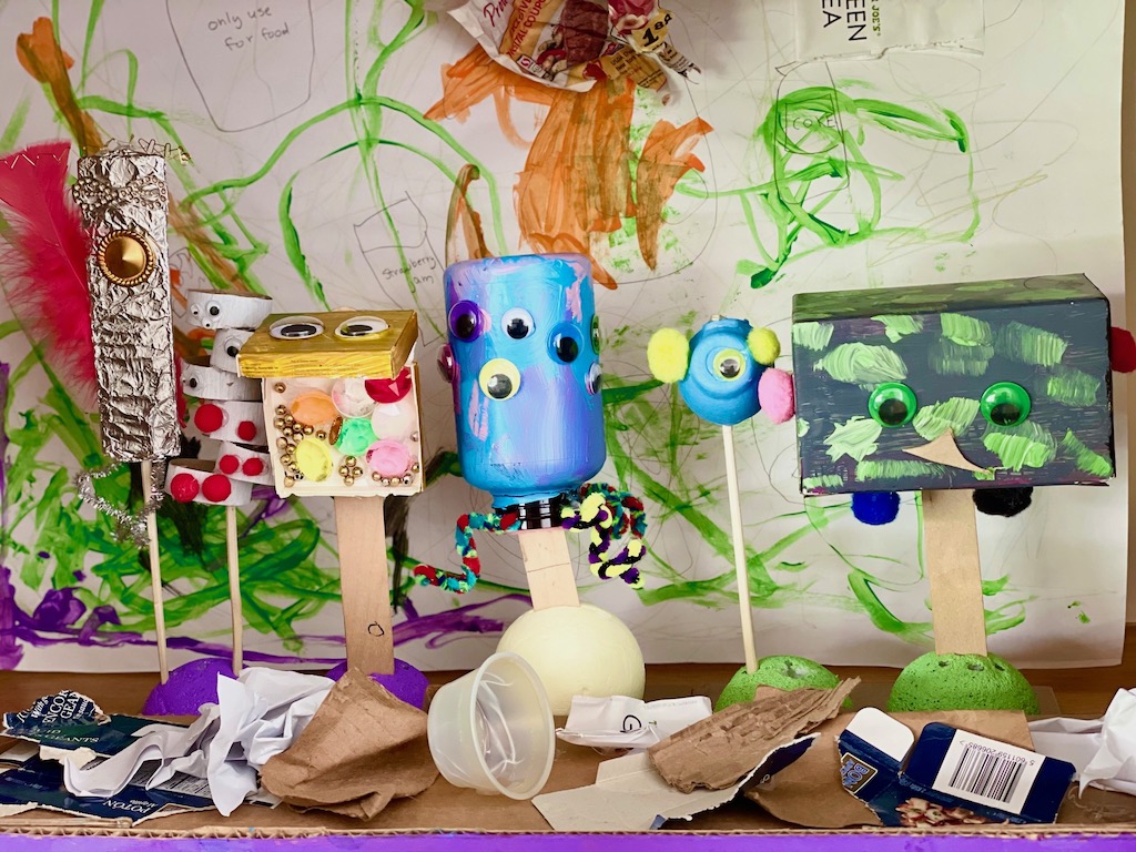 Trash puppets are seen on stage in front of a backdrop of trash and a foreground littered with trash.