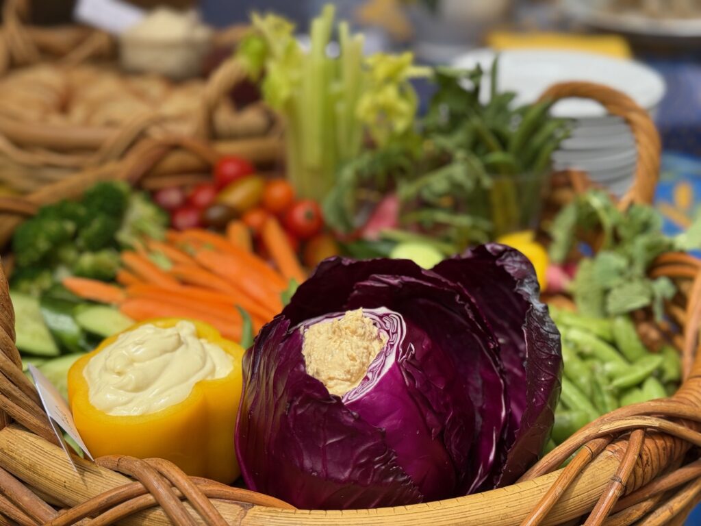 Crudité basket features assorted vegetables and two dips.Aïoli is spooned into a yellow pepper and hummus is served in a hollowed-out purple cabbage.