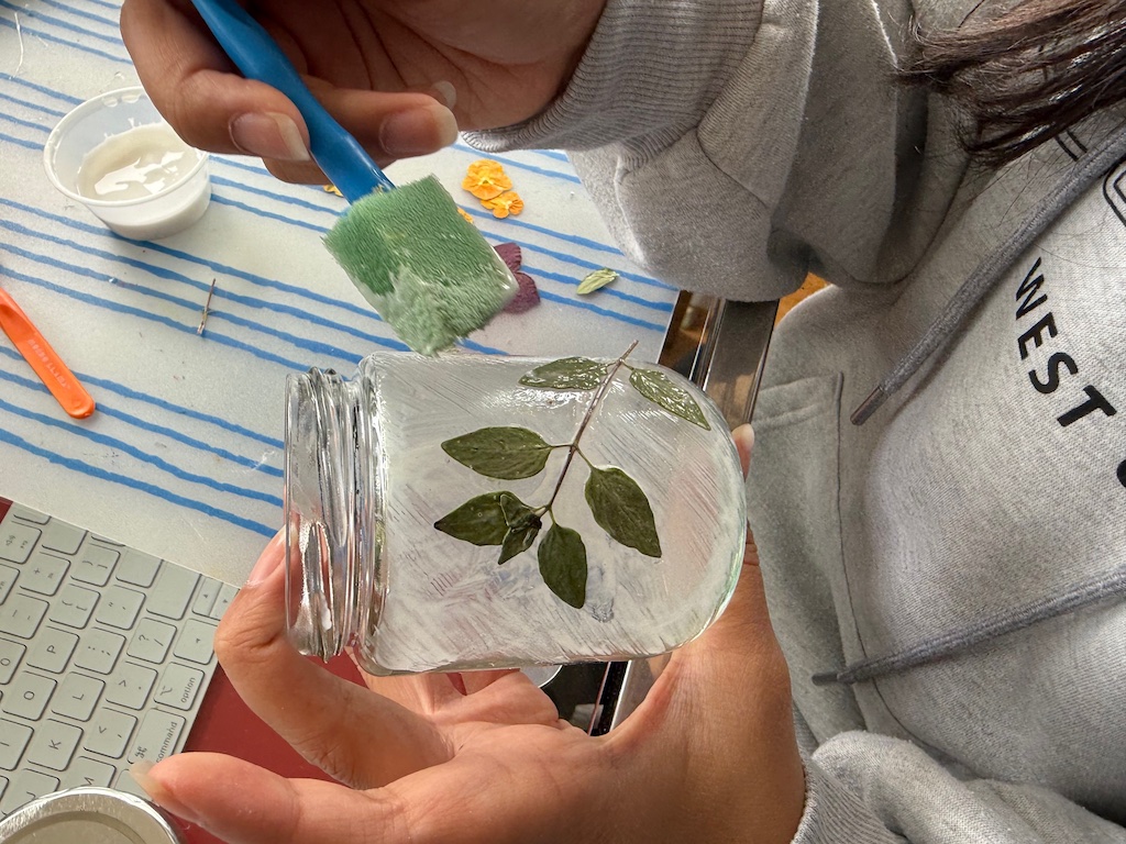 Child brushes white school glue over pressed plant materials and the jar itself to create a translucent finish.