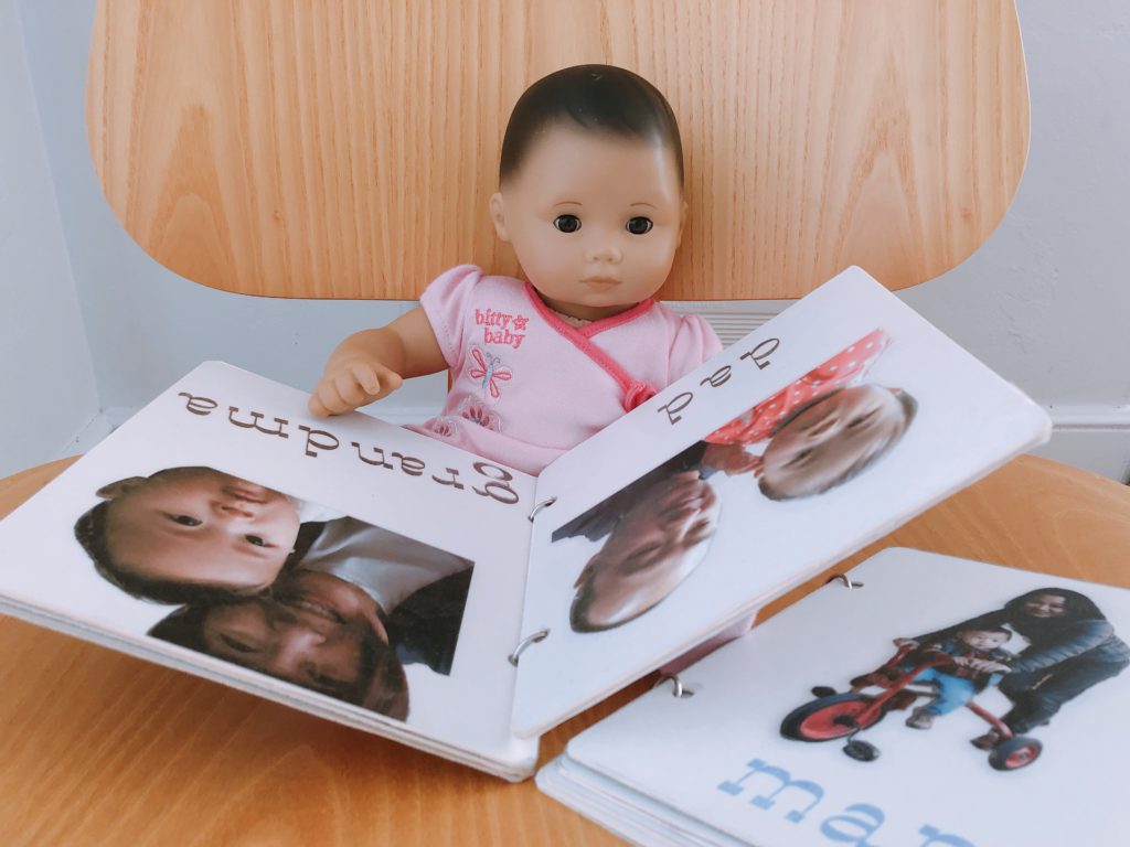 Doll sits with a homemade board book made by a grandma for a toddler. The book is made with cardboard, photos, clear adhesive film, and binder rings.