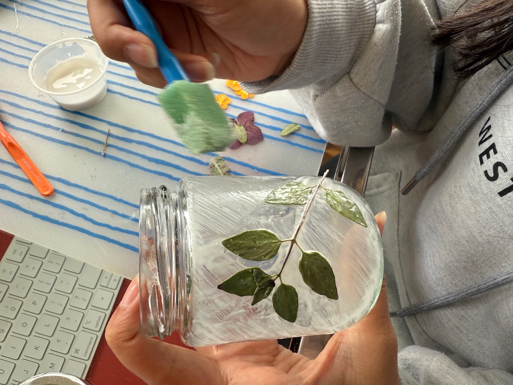 Child paints school glue with a foam brush over pressed flowers on a jar to create a translucent finish.