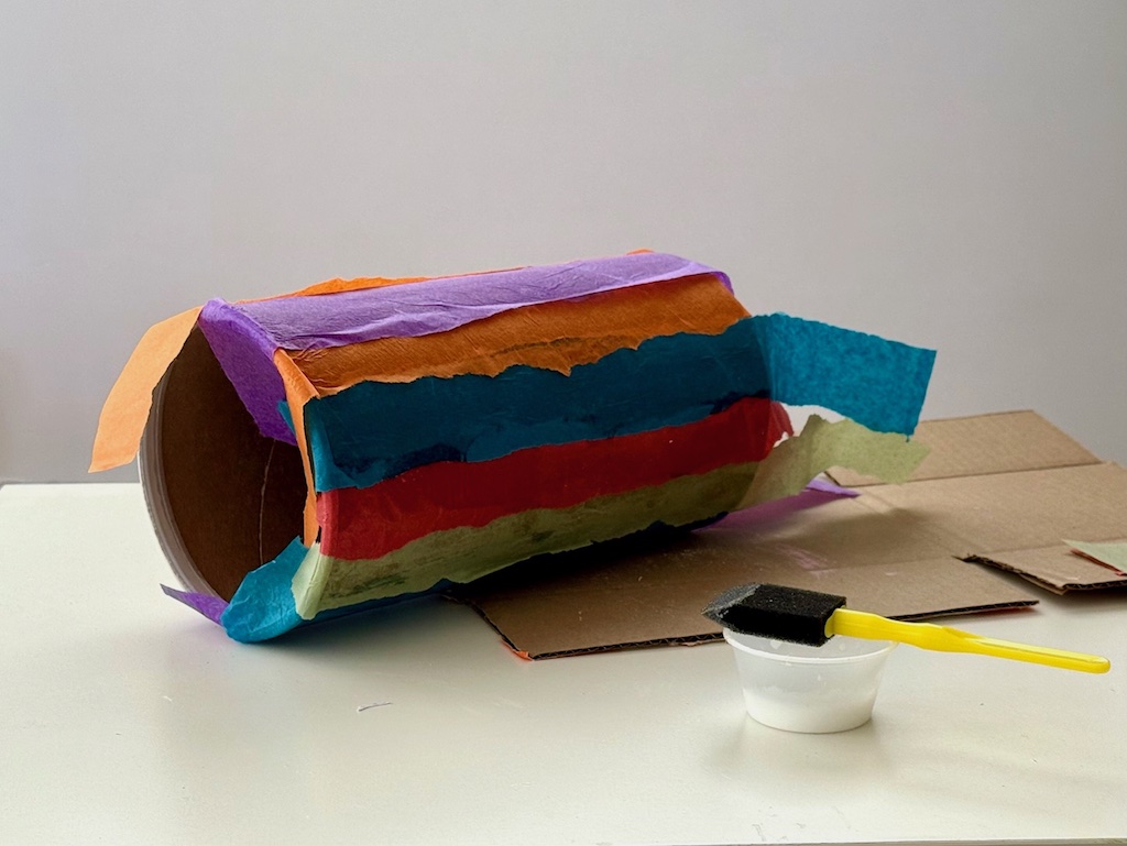 Tissue paper is glued onto the oatmeal container in colorful stripes.