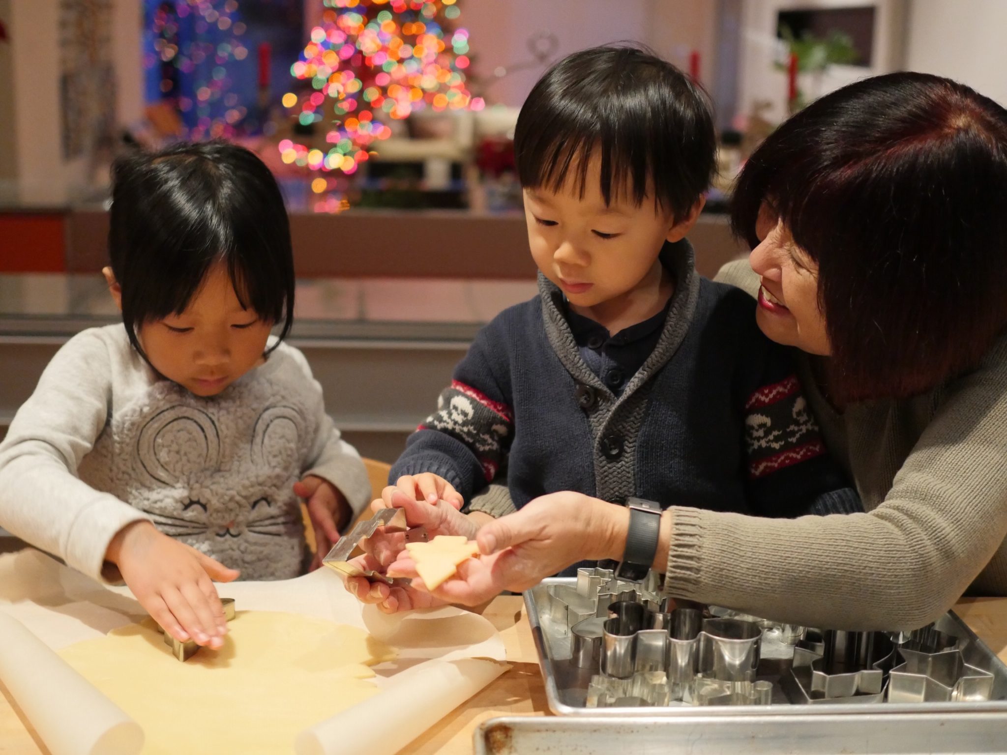 Grandkids and grandma cutting out Christmas cookies with a Christmas tree in the background.