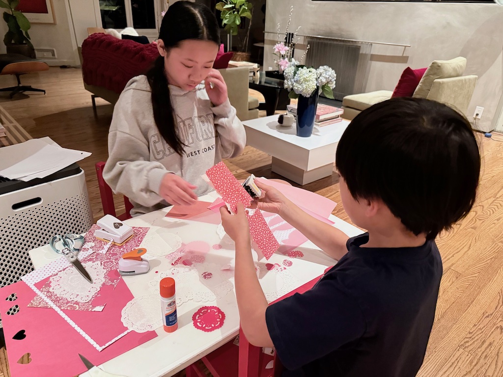 Kids sit at a table to cut out hearts and make valentine placemats. Art supplies include doilies, heart paper punches, and patterned paper.