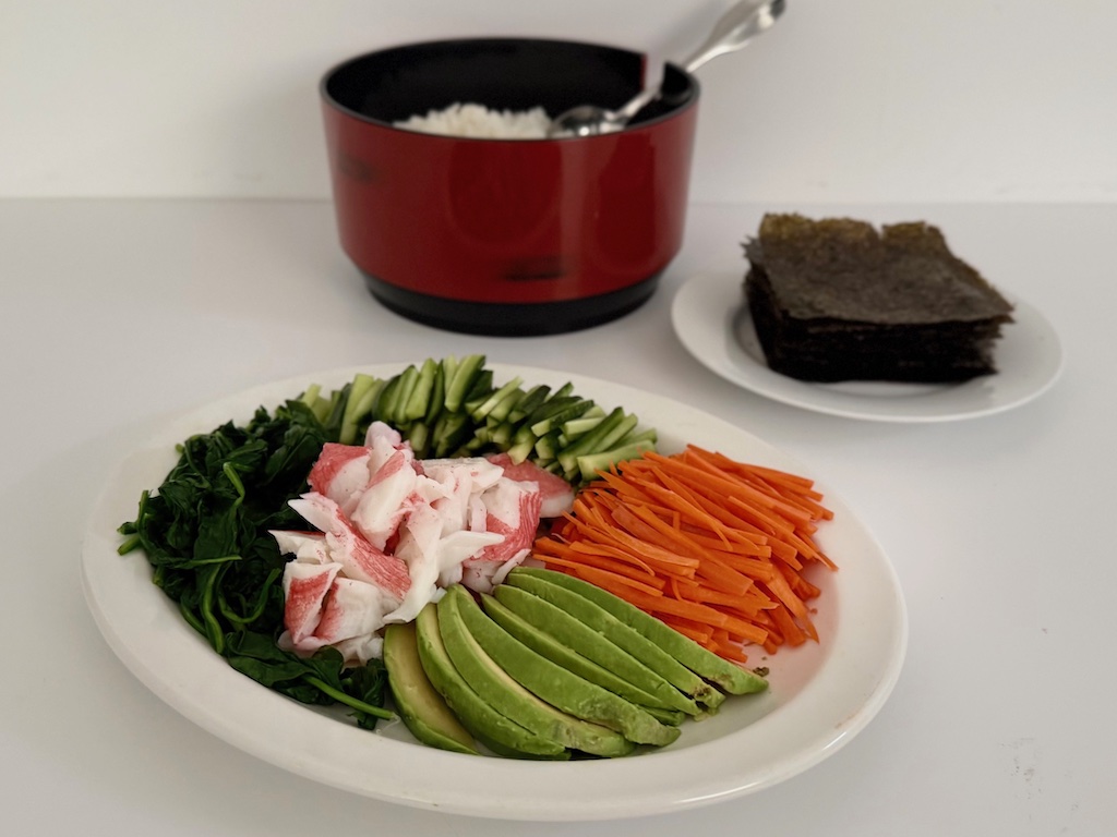 All the sushi fixings: vegetables, and imitation crab are arranged artfully on a platter with nori squares on a plate and seasoned rice in a serving bowl.