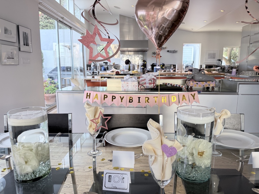 The birthday party table is decorated with hanging ornaments, balloons, and a happy birthday banner. Candle sand centerpieces are set at both sides of the table.