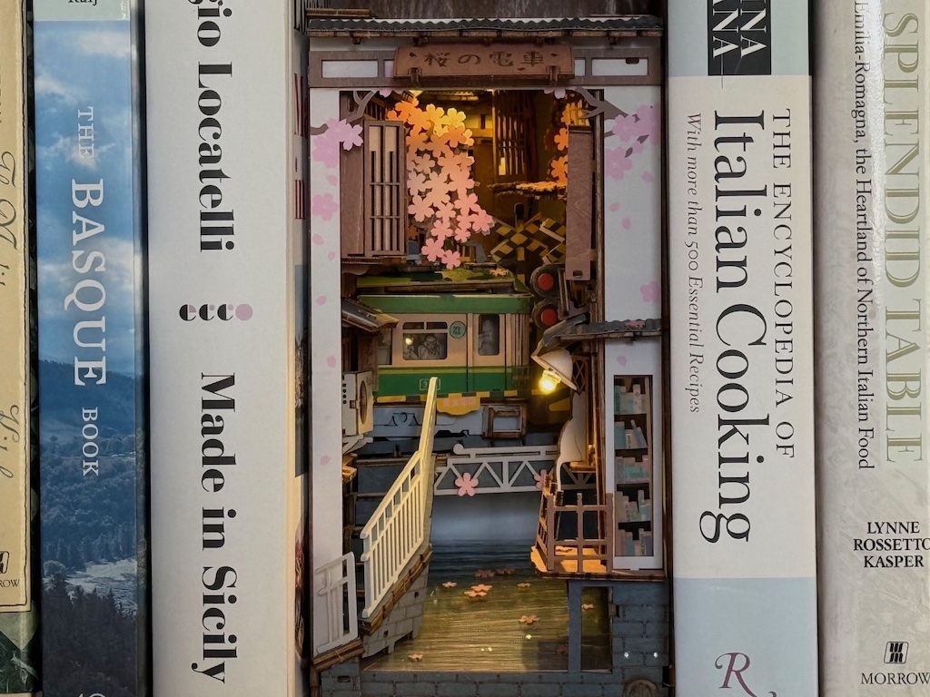 The completed book nook features a Japanese electric train, cherry blossoms, and a stream. The lighted scene sits between two cookbooks on a shelf.