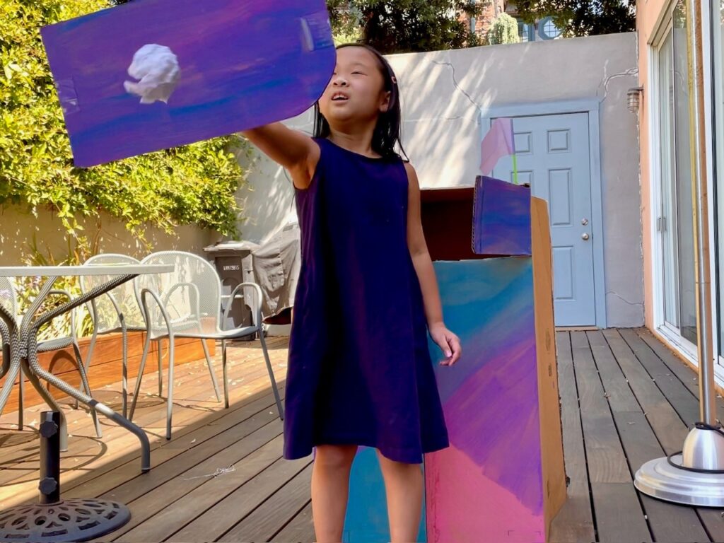 Child uses a cardboard shield to repel paper wad ammunition coming her way. Behind her is a cardboard fort for the fort fight game.