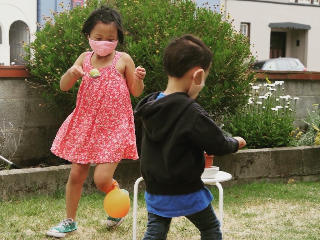 Children, masked during the COVID pandemic, play spoon relay in the grandma's backyard.