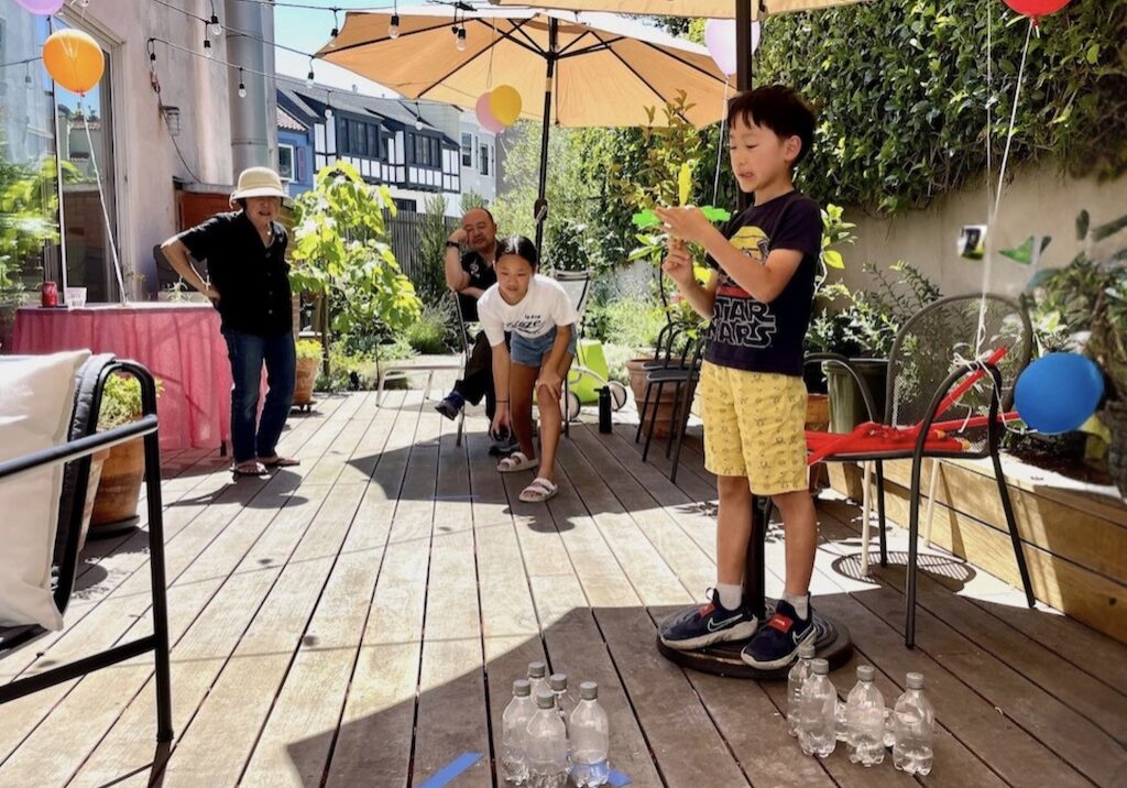 Grandchild tries to knock water bottle pins over during annual summer party.